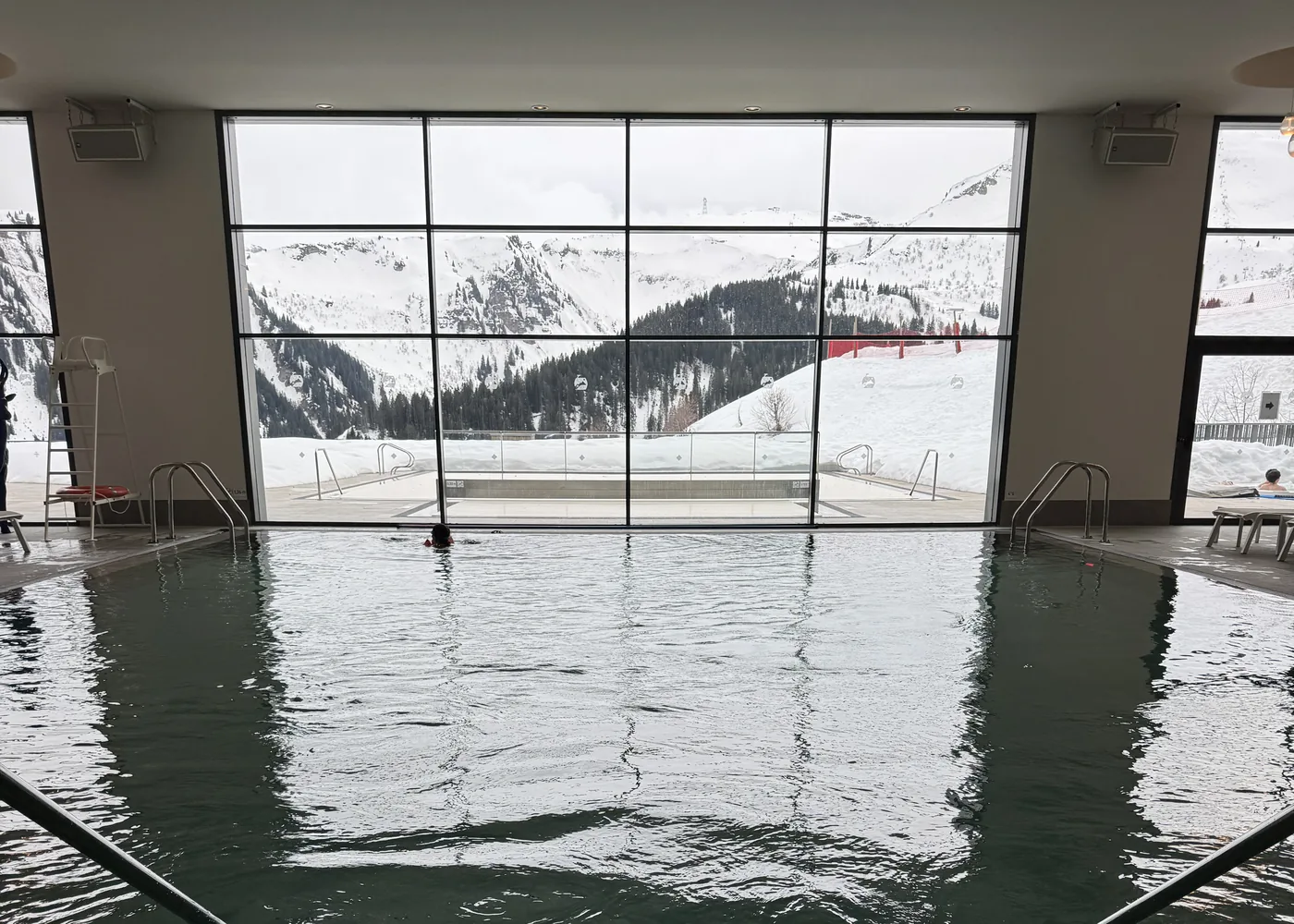Piscina interna aquecida de um resort de neve, com janelão de vidro aberto para as pistas e a montanha nevada
